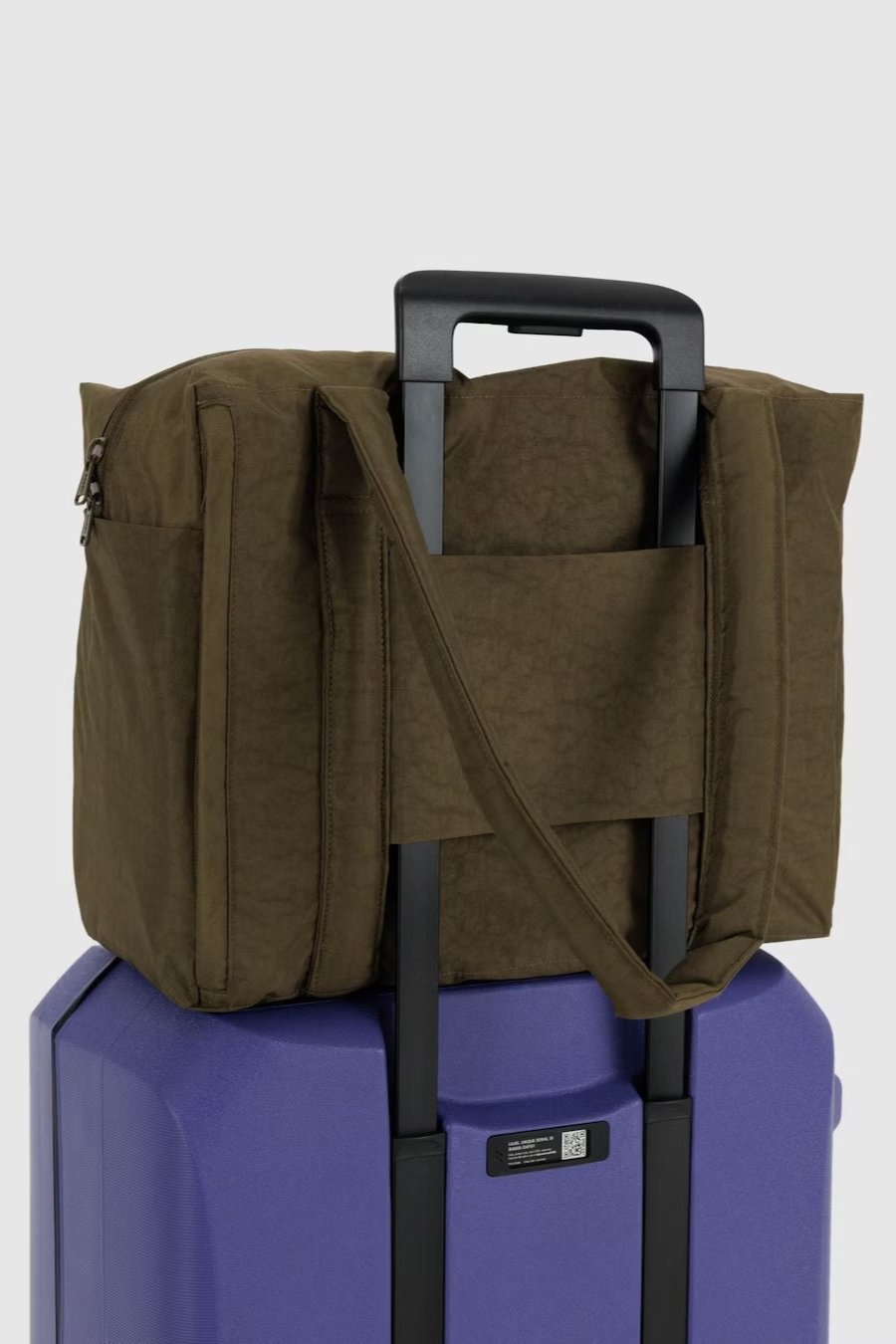 An einem violetten Hartschalenkoffer mit verlängertem Griff ist eine Main Design BAGGU Small Cloud Carry-On Tasche in Olivgrün sicher befestigt, indem man sie über den Griff schiebt. Die Reisetasche verfügt über einen Reißverschluss und Tragegurte. Der Hintergrund ist schlicht weiß.