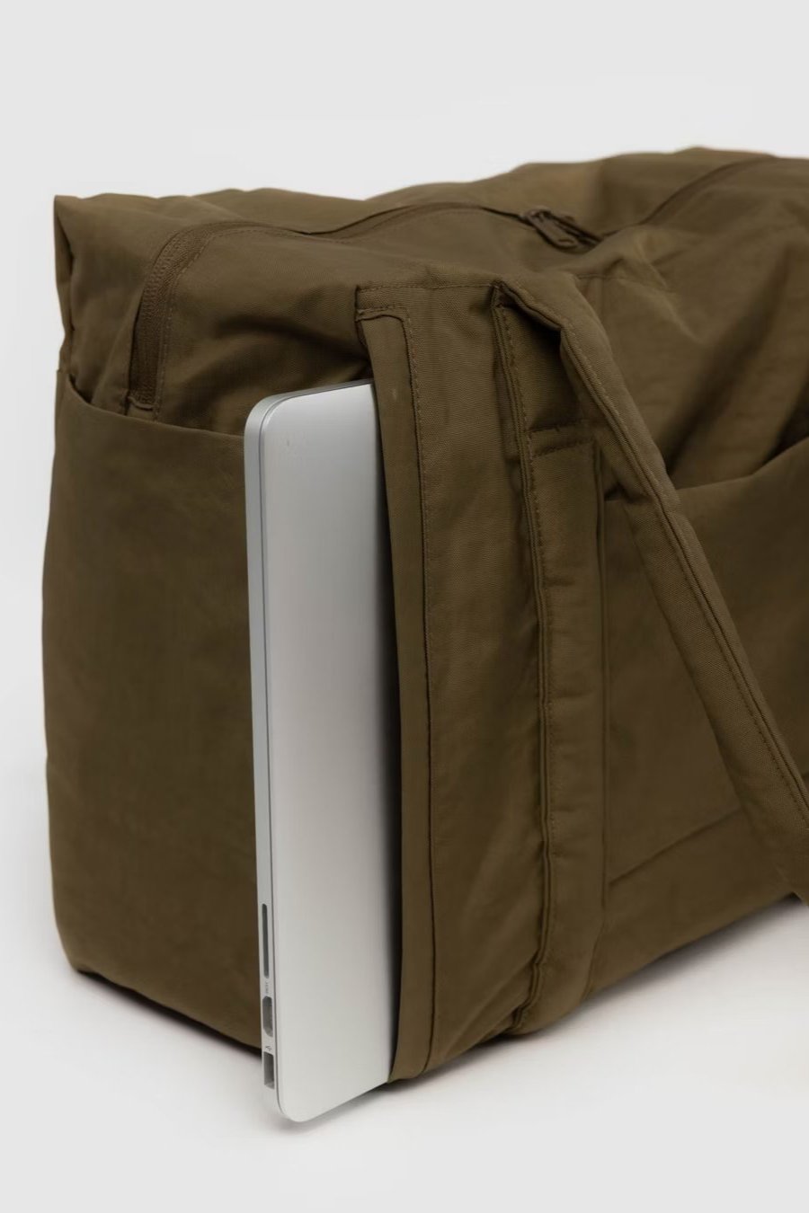 Eine braune BAGGU Small Cloud Carry-On Tasche von Main Design mit einem silbernen Laptop, der teilweise in eine der Seitentaschen gesteckt ist. Die Tasche hat einen Reißverschluss und einen Schultergurt und steht vor einem schlichten, hellen Hintergrund.