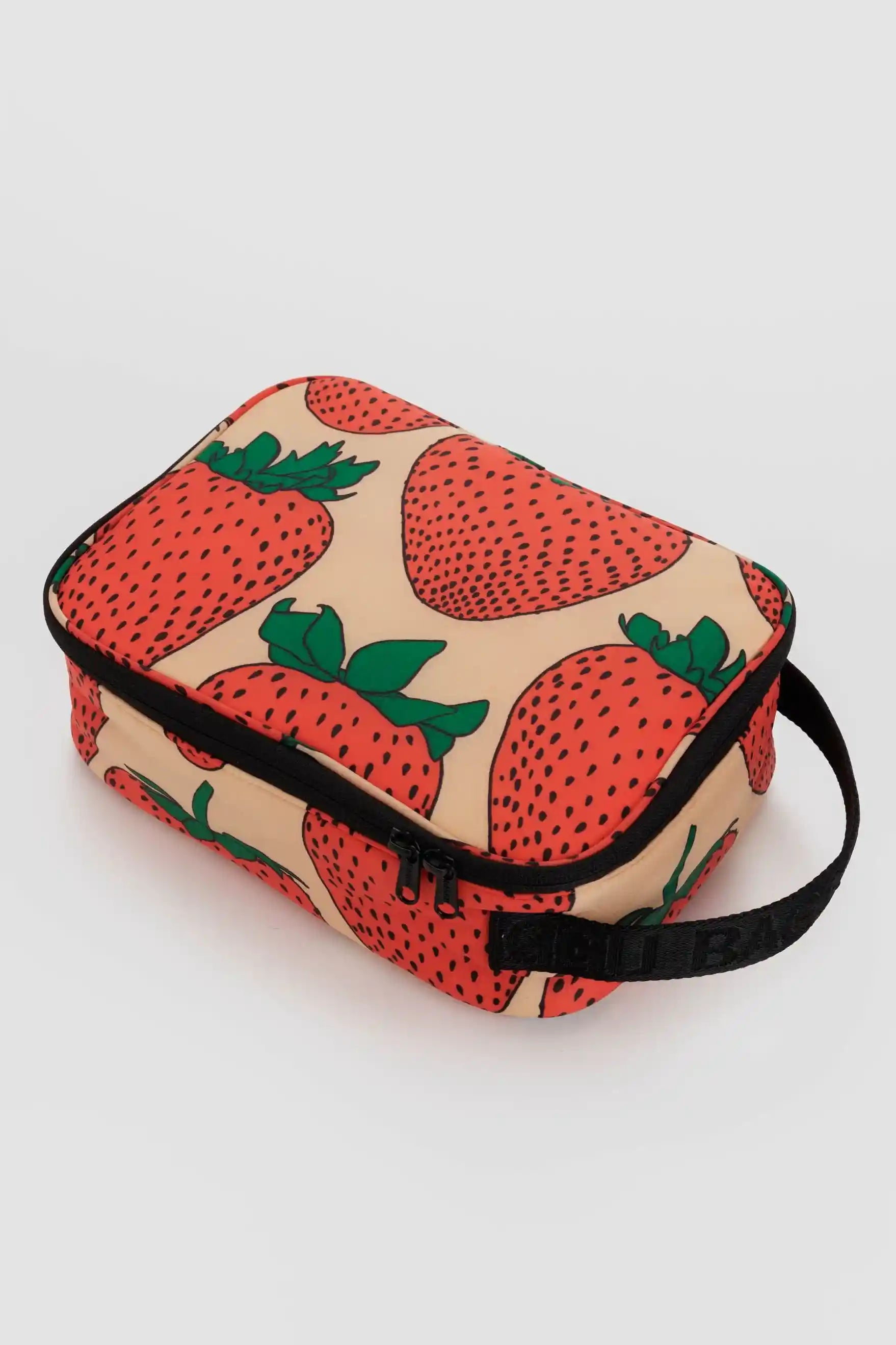 Die BAGGU Lunch Box von Main Design ist eine rechteckige, beigefarbene Kosmetiktasche aus recyceltem Nylon mit einem lebendigen, fruchtigen Erdbeermuster. Sie hat einen schwarzen Reißverschluss an der Oberseite und einen schwarzen Riemen an einer Seite zum einfachen Tragen. Die Erdbeeren sind rot mit grünen Blättern und verleihen dem Design einen fröhlichen Farbtupfer.