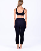 Eine Frau mit langen Haaren steht barfuß und blickt von der Kamera weg. Sie trägt einen schwarzen Sport-BH und hoch taillierte Bahamas-Leggings von Main Design aus nachhaltigem Stoff. Der Hintergrund ist schlicht weiß.