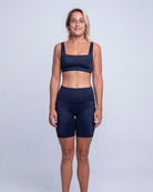 Eine Frau mit schulterlangem blondem Haar steht vor einem schlichten Hintergrund und trägt das Betty Active Top von Main Design, einen schwarzen Sport-BH mit breiten Trägern und schwarze Sportshorts aus recycelten Materialien. Sie ist barfuß und hat einen entspannten, neutralen Gesichtsausdruck, ihre Arme ruhen an ihren Seiten.