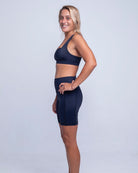 Eine Person steht barfuß vor einem weißen Hintergrund und blickt nach links. Sie trägt ein marineblaues Betty Active Top von Main Design und dazu passende hoch taillierte Radlerhosen aus recyceltem Material. Die Person hat blonde Haare, lächelt und ihre Arme hängen bequem an den Seiten.