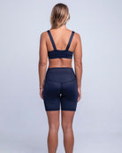 Eine Person mit langen Haaren steht barfuß vor einem einfarbigen Hintergrund und blickt von der Kamera weg. Sie trägt ein dunkles, ärmelloses Betty Active Top von Main Design und dazu passende High-Waist-Shorts aus recycelten Materialien, die Arme hängen an den Seiten.