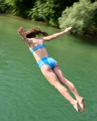 Eine Frau in einem hellblauen Sassy Bikini Oberteil reversibel von Main Design springt mitten in der Luft in einen grünen Fluss, mit Bäumen und Büschen entlang des Ufers im Hintergrund.