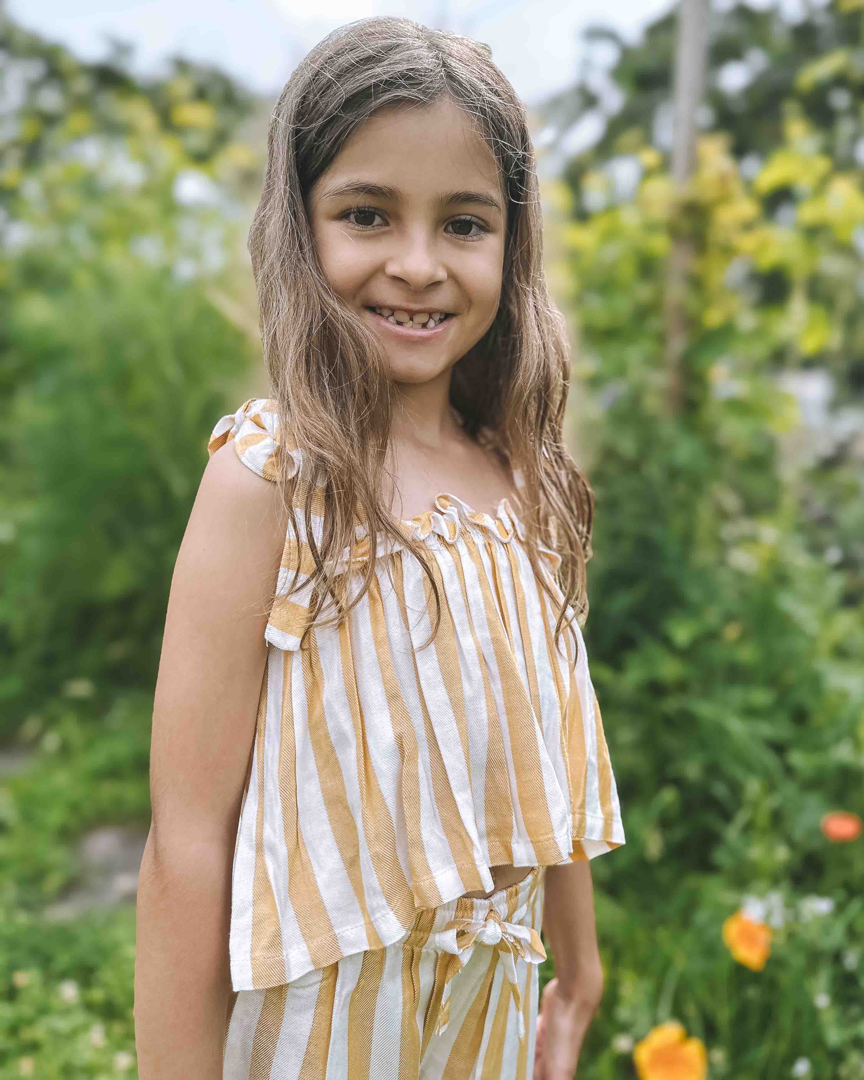 Lächelndes junges Mädchen mit langen braunen Haaren trägt das Main Design Boho Hawaii Set, ein gelb-weiß gestreiftes Outfit, während sie draußen in einem üppigen grünen Garten mit orangefarbenen Blumen und verschwommenem Laub im Hintergrund steht.