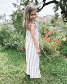 Ein junges Mädchen mit langen braunen Haaren, das den ärmellosen, gestreiften Overall „Boho Hawaii Set“ von Main Design trägt, steht lächelnd auf dem Gras in einem Garten mit grünen Pflanzen und roten Blumen hinter ihr.