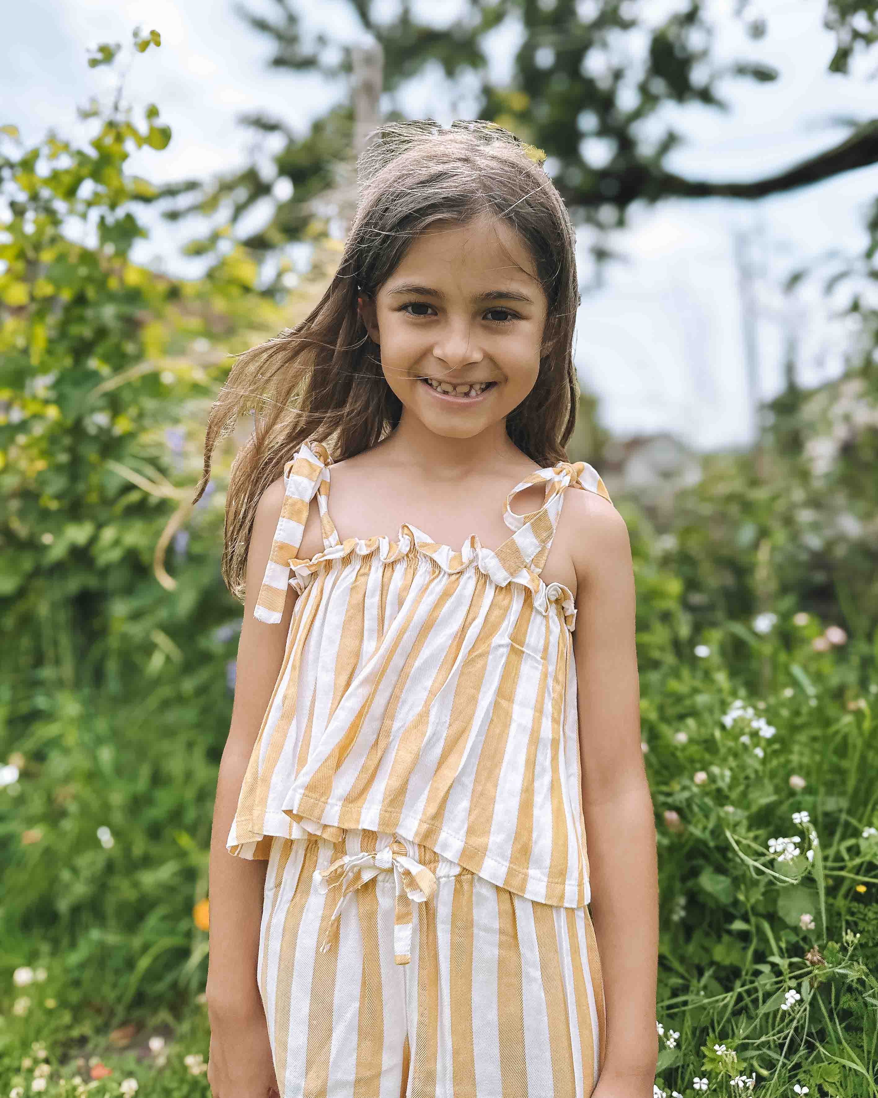 Ein junges Mädchen mit langen braunen Haaren lächelt draußen in einem grünen, blumenreichen Garten und trägt das Boho Hawaii Set von Main Design.