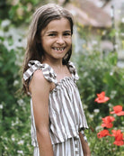 Ein junges Mädchen mit langen braunen Haaren lächelt im Freien. Es trägt das Main Design Boho Hawaii Set – einen ärmellosen, gestreiften Mädchen-Zweiteiler mit gebundenen Trägern. Sie steht in einem Garten, umgeben von grünem Laub und roten Blumen.