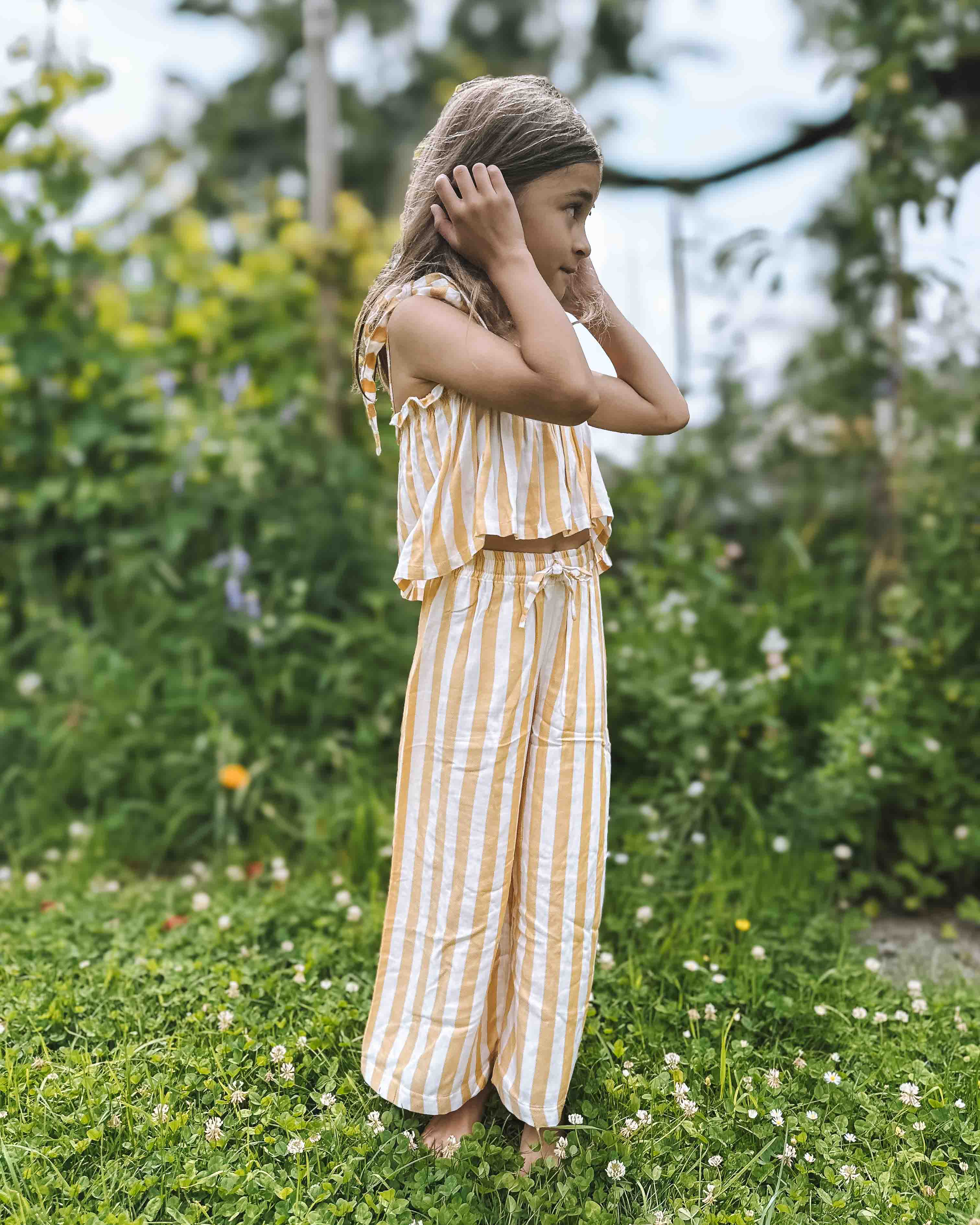 Ein junges Mädchen mit hellbraunem Haar steht barfuß im Gras eines Gartens und trägt das Boho Hawaii Set von Main Design. Sie hält ihre Hände an den Ohren und blickt zur Seite vor einem Hintergrund aus grünen, blattreichen Pflanzen.