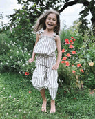 Ein lächelndes junges Mädchen mit langen Haaren steht barfuß auf dem Gras in einem Garten und trägt das Boho Hawaii Set von Main Design, mit grünen Pflanzen und roten Blumen im Hintergrund.