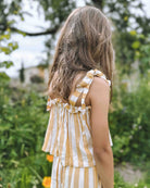 Ein junges Mädchen mit hellbraunem Haar steht im Freien und trägt das Boho Hawaii Set von Main Design mit gelben und weißen Streifen, umgeben von üppigem grünem Laub und verschwommenen Blumen.