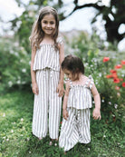 Zwei kleine Kinder stehen draußen auf der Wiese. Beide tragen passende hell gestreifte Outfits aus dem Main Design Boho Hawaii Set. Das ältere Mädchen lächelt in die Kamera, während leuchtende Blumen und Grün sie in ihrem stilvollen Set umgeben.