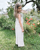 Ein junges Mädchen mit langen braunen Haaren, das den ärmellosen, gestreiften Overall „Boho Hawaii Set“ von Main Design trägt, steht lächelnd auf dem Gras in einem Garten mit grünen Pflanzen und roten Blumen hinter ihr.