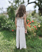 Ein junges Mädchen mit langen braunen Haaren, gekleidet im Boho Hawaii Set von Main Design, steht mit dem Rücken zur Kamera in einem Garten voller grünem Gras und roten Blumen.