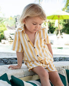 Ein junges blondes Kind sitzt auf einem Steinvorsprung neben einem Pool und trägt das Boho-Shirt-Set von Main Design – stylische Sommerkleidung für Kinder, ideal für sonnige Tage.