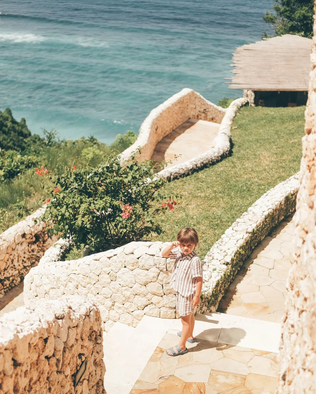 Ein kleines Kind trägt das Boho-Shirt-Set von Main Design und steht auf einem von weißen Mauern und Grün gesäumten Steinpfad mit Blick auf türkisfarbene Meereswellen in einer sonnigen, friedlichen Umgebung mit üppigem Gras und blühenden Büschen.