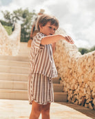 Ein kleines Kind in einem Boho-Shirt-Set von Main Design, ideal für Kinder-Sommerkleidung, steht mit ausgestrecktem Arm auf sonnigen Steinstufen. Dahinter sind Steinmauern und Grünflächen unter einem teilweise bewölkten Himmel zu sehen.