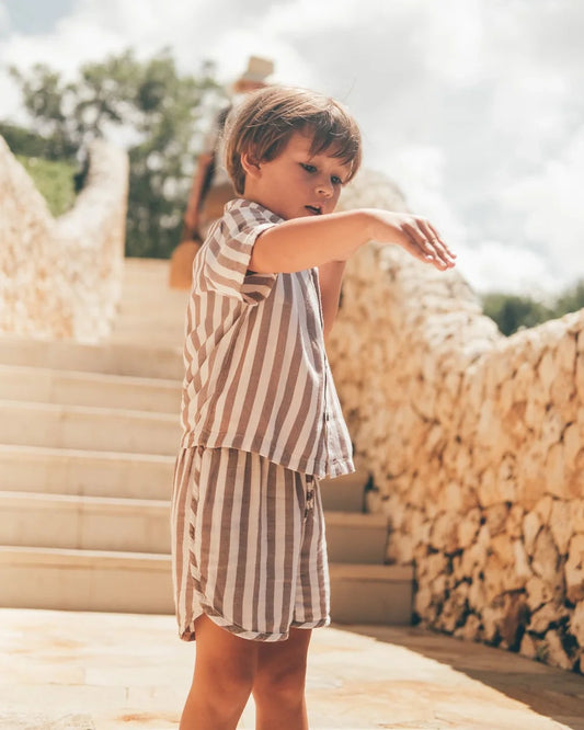 Ein kleines Kind in einem Boho-Shirt-Set von Main Design, ideal für Kinder-Sommerkleidung, steht mit ausgestrecktem Arm auf sonnigen Steinstufen. Dahinter sind Steinmauern und Grünflächen unter einem teilweise bewölkten Himmel zu sehen.