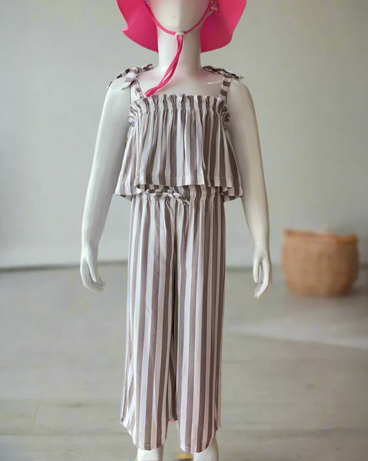 Eine Kinderpuppe präsentiert das Boho Hawaii Set von Main Design: einen ärmellosen, gerüschten, grau-weiß gestreiften Overall mit gebundenen Schulterträgern und weiten Beinen, darüber ein leuchtend rosa Sonnenhut für einen perfekten Sommertraum-Stil.