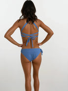 Eine Frau blickt von der Kamera ab. Sie trägt das Main Design Charm Surf Bikini Oberteil in Blau mit Bindedetails aus chlorresistentem Stoff. Ihre Hände sind auf den Hüften gestemmt. Ihr schulterlanges braunes Haar hebt sich von einem schlichten hellen Hintergrund ab.