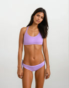 Eine Frau mit langen dunklen Haaren trägt das Charm Surf Bikini Oberteil von Main Design in Hellviolett vor schlichtem Hintergrund und steht entspannt da – ideal für Wassersport dank UPF 50+ Sonnenschutz.