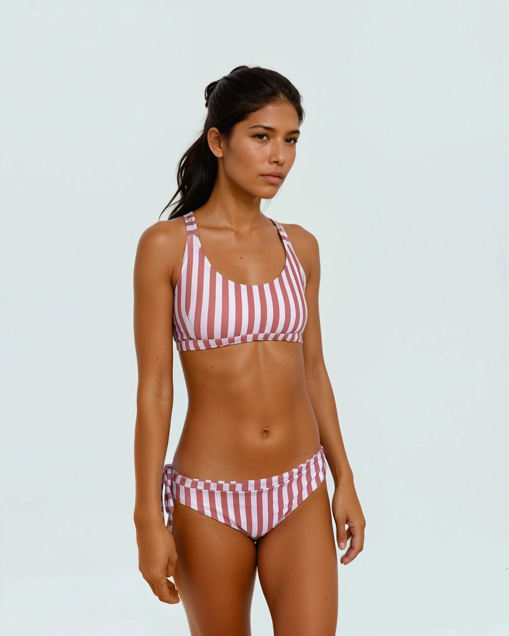 Eine Frau trägt im Freien die Main Design Blush Surf Bikinihose mit rosa und weißen Streifen mit neutralem Gesichtsausdruck vor einem schlichten hellen Hintergrund.
