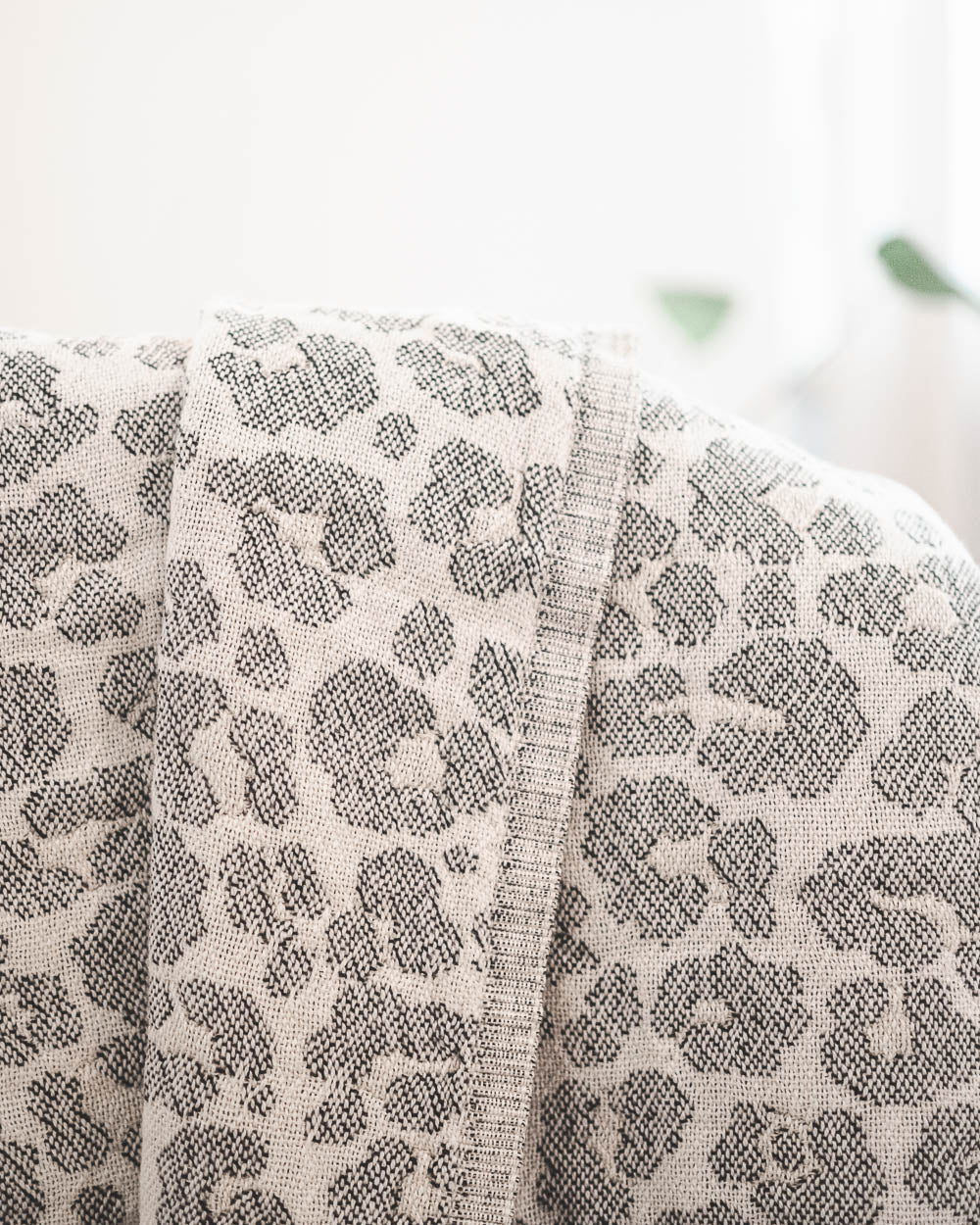 Eine Nahaufnahme der gefalteten, hellen Decke Savanna von Main Design mit grauem Leopardenmuster, drapiert auf einer Fläche mit sanft verschwommenem Hintergrund.