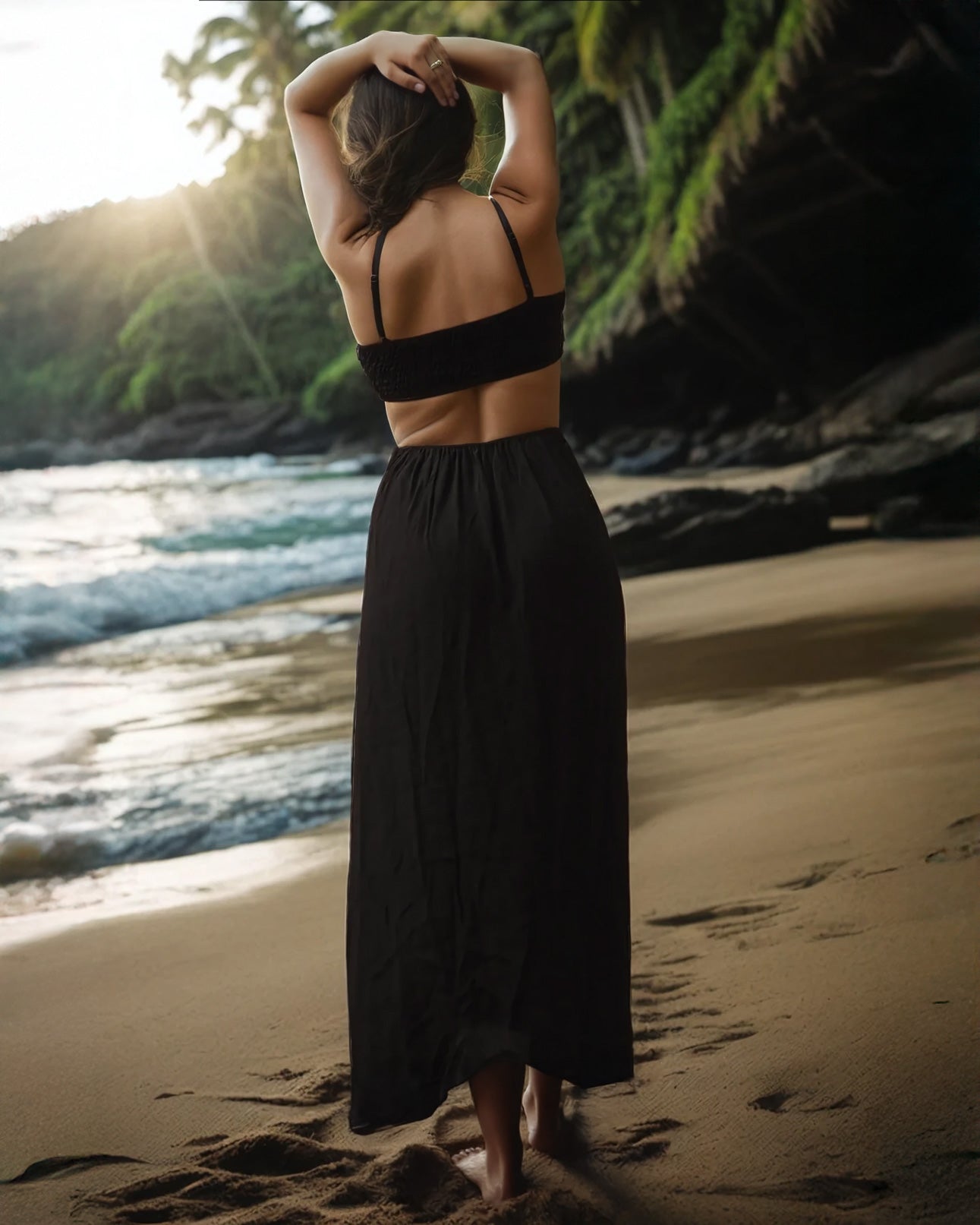 Eine Frau im Maxikleid Chasing the Sun von Main Design steht an einem Sandstrand und streckt die Arme zum Meer aus. Die untergehende Sonne, teilweise von Palmen verdeckt, wirft einen warmen Schein, während die Wellen sanft ans Ufer plätschern.