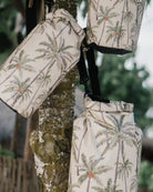 Drei Dry Bags von Meraki Supply Co. von Main Design, jede mit einem tropischen Palmenmuster, hängen an einem Baumstamm. Der Hintergrund besteht aus Grünpflanzen und einem Holzzaun, während jede Tasche über einen abnehmbaren Schultergurt für zusätzliche Vielseitigkeit verfügt.