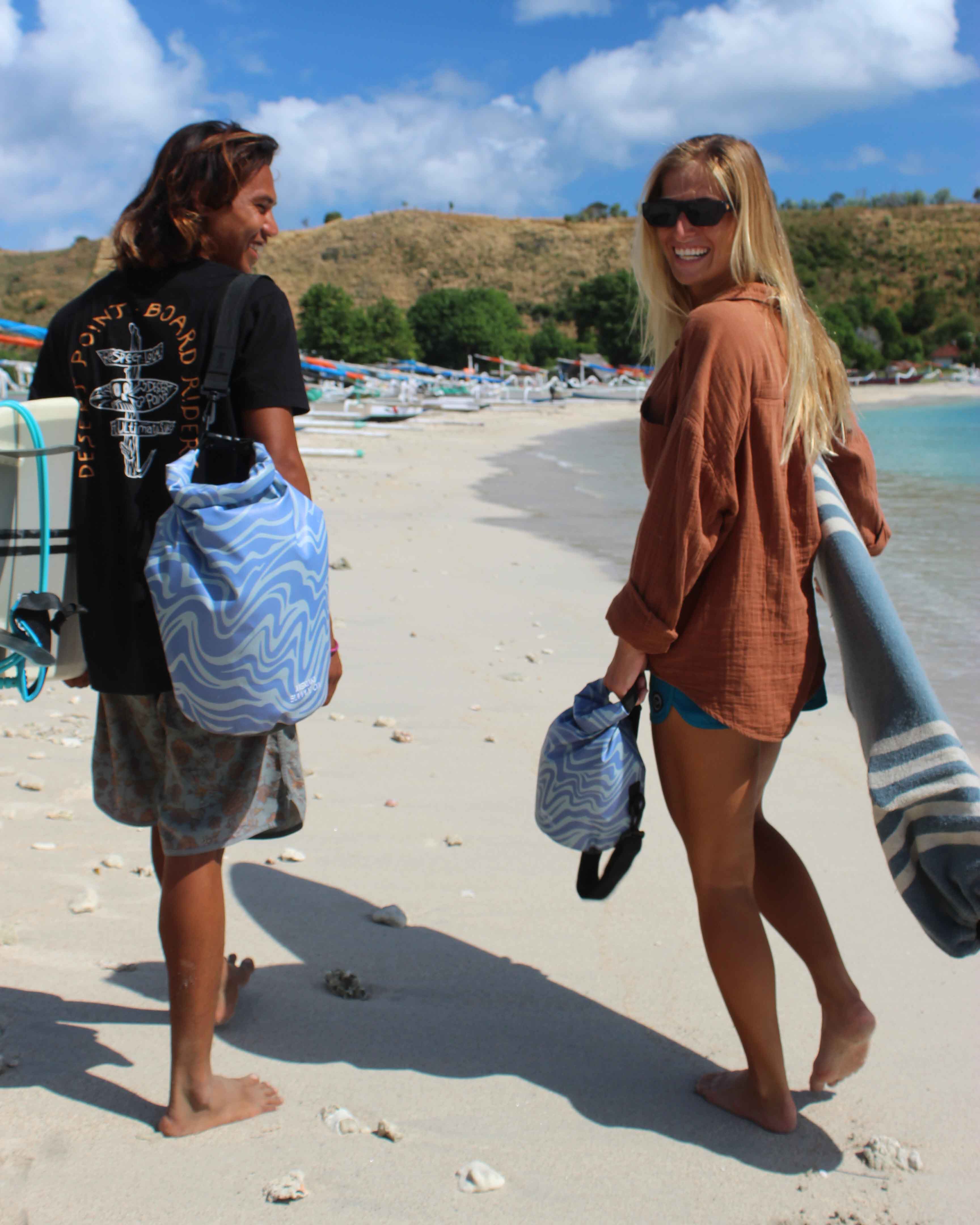 Ein lächelnder Mann und eine lächelnde Frau laufen barfuß mit Surfbrettern, Handtüchern und einem Dry Bag von Meraki Supply Co. von Main Design an einem Sandstrand entlang. Boote und Bäume prägen den Hintergrund unter einem klaren blauen Himmel.