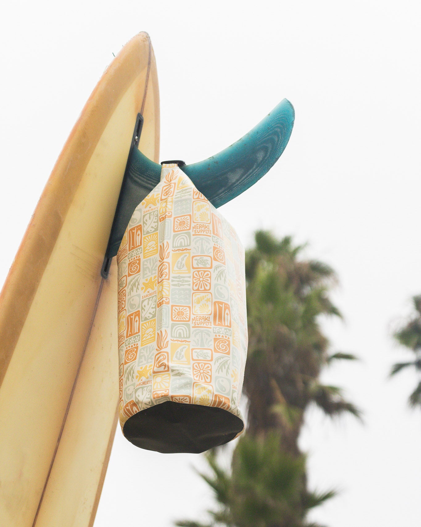 Die Dry Bag von Meraki Supply Co. by Main Design mit orangefarbenen und blaugrünen Mustern hängt an einer Surfbrettflosse mit Palmen und einem bewölkten Himmel im Hintergrund.