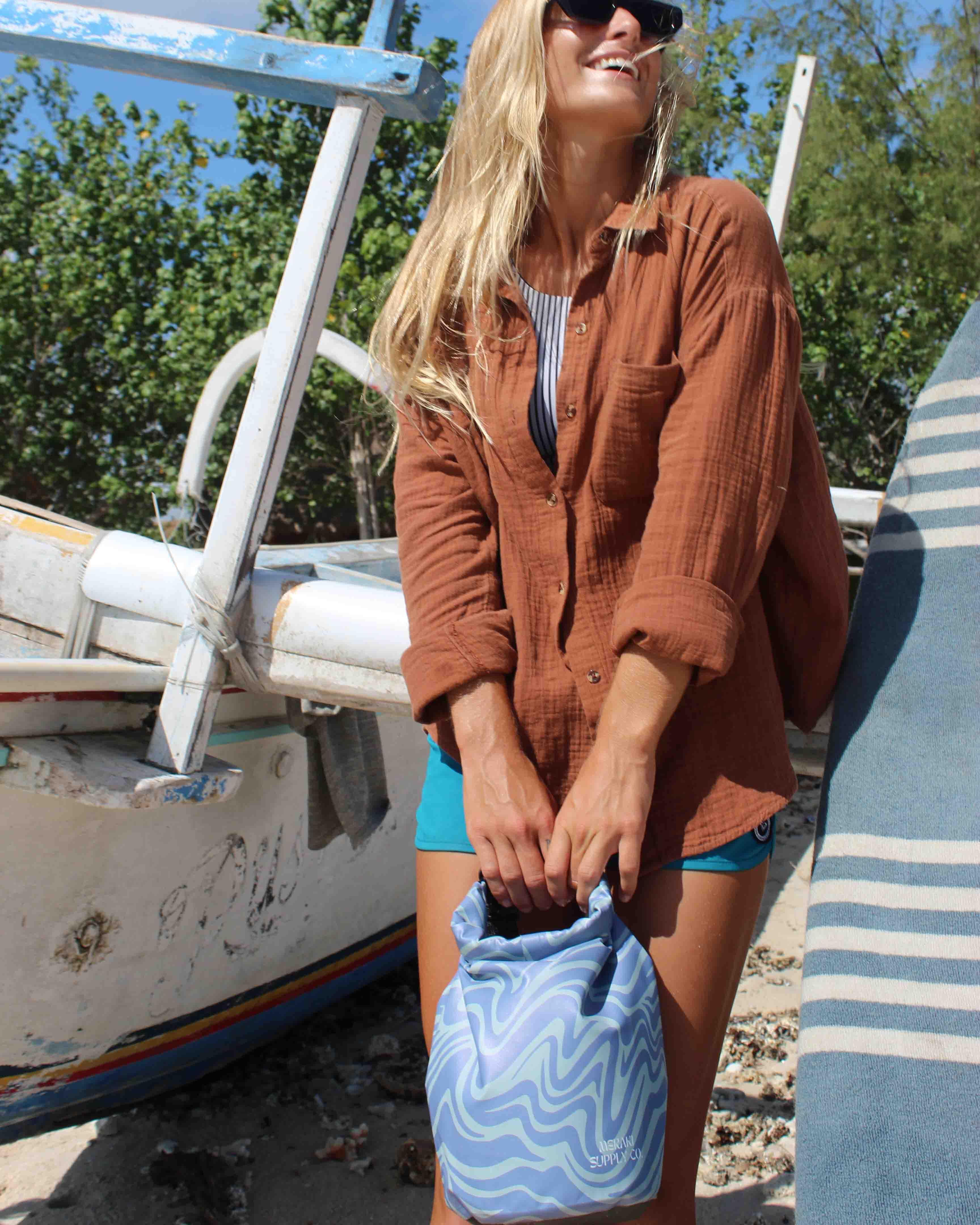 Eine blonde Frau mit Sonnenbrille, braunem Hemd und blauen Shorts lächelt am Strand neben einem Boot und hält eine Main Design Dry Bag von Meraki Supply Co. in der Hand; im Hintergrund sind Bäume und ein gestreiftes Handtuch zu sehen.