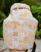 Eine Nahaufnahme eines farbenfrohen, wasserdichten Dry Bag von Meraki Supply Co. von Main Design mit von der Natur inspirierten Mustern und Markenzeichen vor üppigen grünen tropischen Pflanzen.