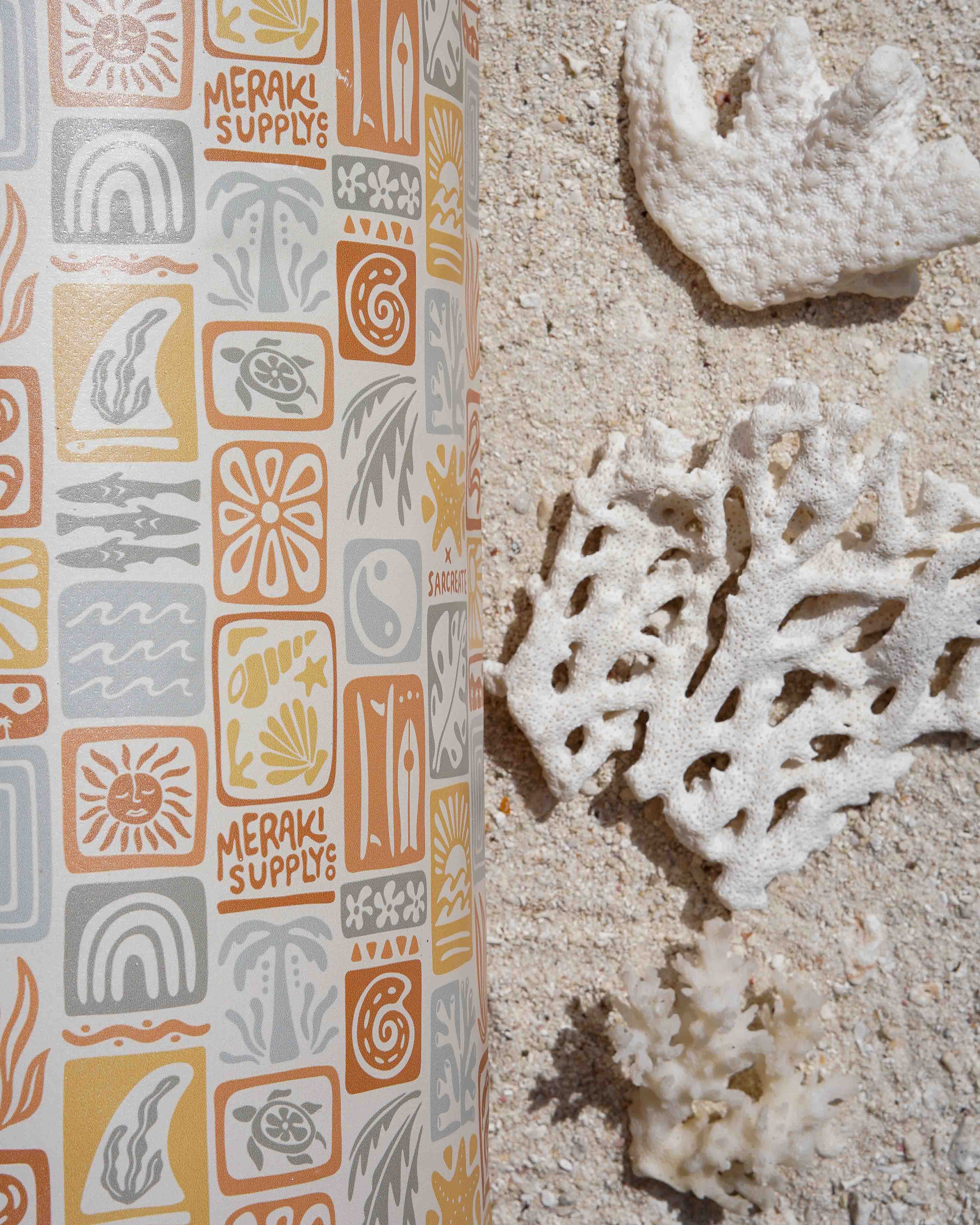 Eine Yogamatte mit orangefarbenen, gelben und grauen Mustern und dem Text „MERAKI SUPPLY CO.“ liegt auf sandigem Boden neben weißen Korallen – perfekt in Kombination mit dem wasserdichten Dry Bag von Meraki Supply Co. von Main Design für Ihre Strandabenteuer.