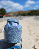 Ein Dry Bag von Meraki Supply Co. by Main Design mit Wellenmuster und wasserdichtem Material liegt an einem Sandstrand in der Nähe eines blauen Seils mit verschwommenen Bäumen, Booten und Hügeln im teilweise bewölkten Hintergrund.