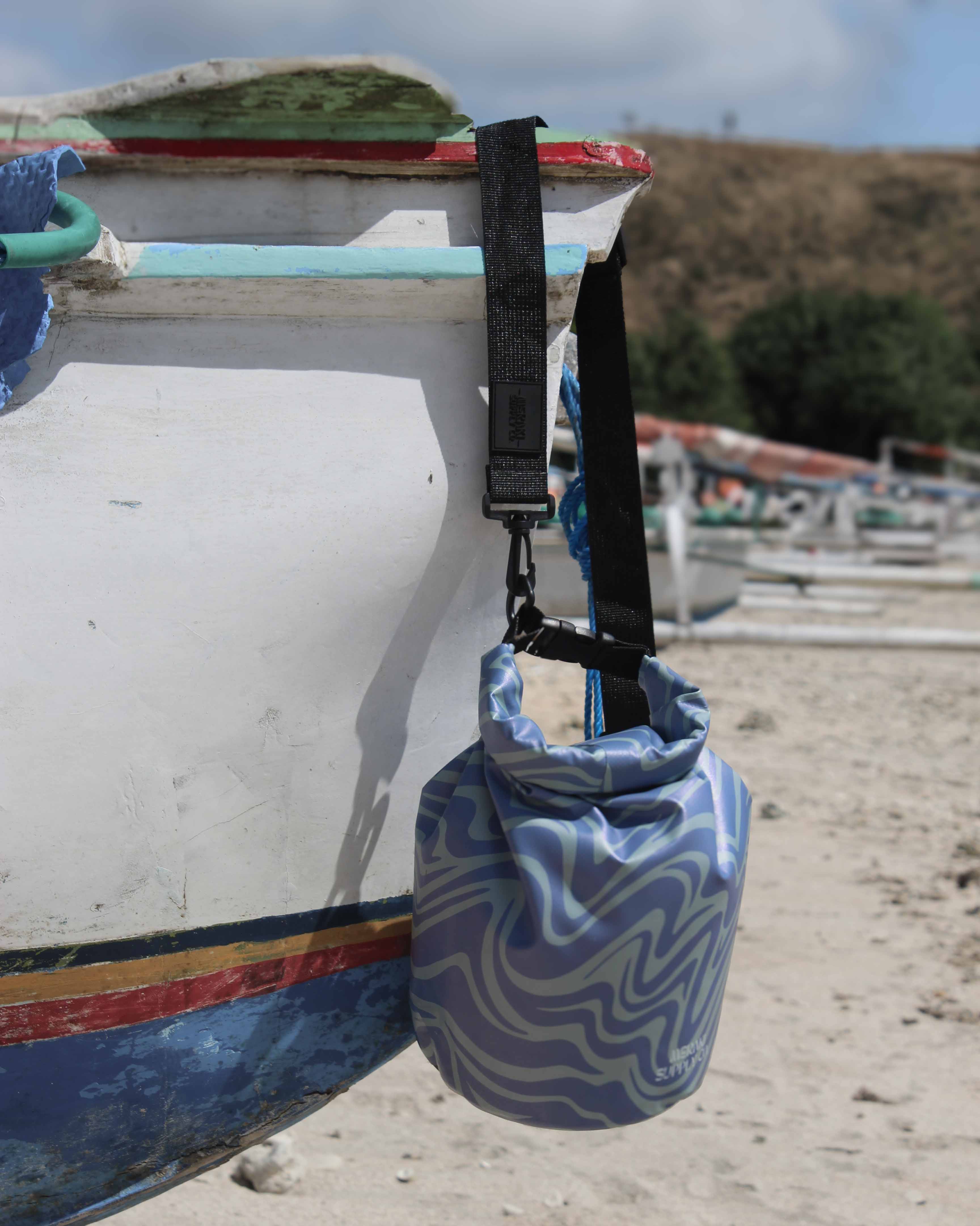 Eine kleine blau-weiße Dry Bag mit Wellenmuster von Meraki Supply Co. by Main Design hängt an einem schwarzen Riemen an einem Holzboot am Strand, mit Hügeln und anderen Booten dahinter, und hält Ihre Ausrüstung wasserdicht.