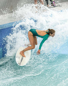In einem grünen Main Design Eclipse Surf Suit surft eine Frau auf einer künstlichen Welle, geduckt auf ihrem Brett inmitten von plätscherndem Wasser, während im Hintergrund andere Surfer und Bretter zu sehen sind.