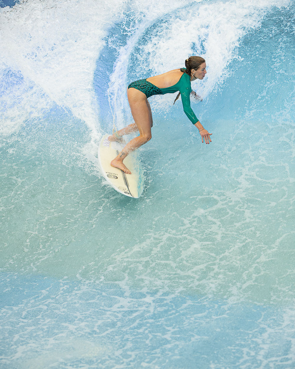 Eine Frau, die den aus nachhaltigem Material hergestellten Eclipse Surf Suit von Main Design trägt, surft auf einer türkisfarbenen Welle auf einem weißen Brett, während das Wasser hinter ihr aufspritzt.
