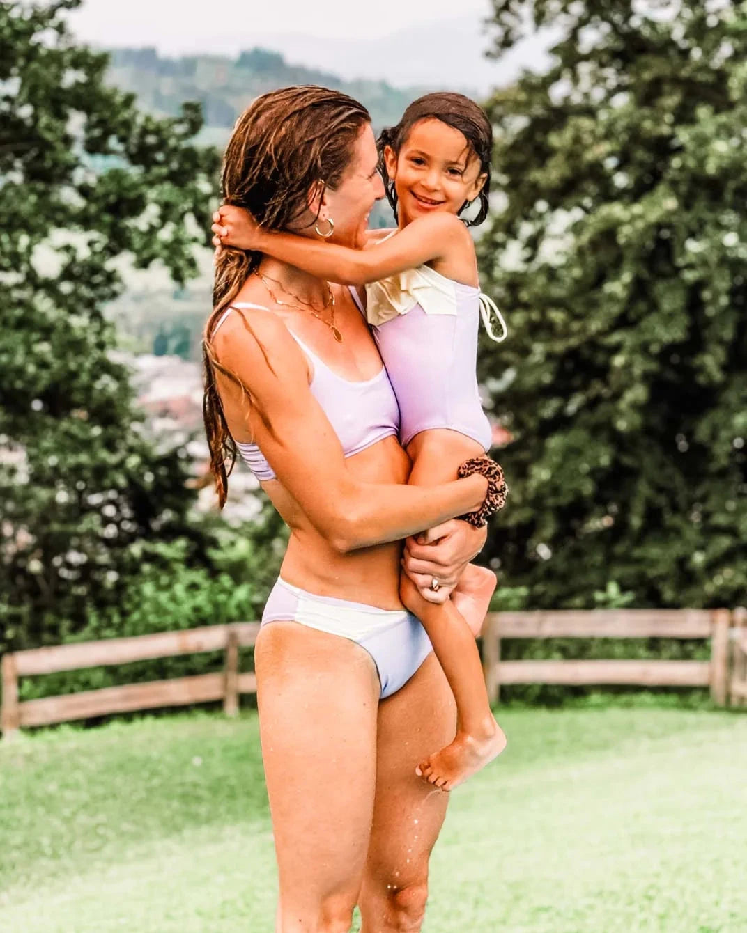 Eine Frau in einem hellvioletten Badeanzug aus der Cali Coast Collection hält ein lächelndes Kind, das Main Designs Flair Bikinihose in einem Farbblockmuster aus Violett und Gelb trägt. Sie stehen auf grünem Gras mit Bäumen und einer verschwommenen Landschaft im Hintergrund und wirken glücklich und entspannt.