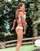 Eine Frau in einer Main Design Flair Bikinihose steht am Beckenrand und hält ein kleines Kind im Arm. Beide sind nass, was darauf schließen lässt, dass sie schwimmen waren. Die Szene spielt vor einem Hintergrund aus Gras, Bäumen und einem Zaun.