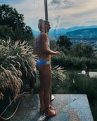 Eine Person, die die Main Design Flair Bikinihose trägt, duscht im Freien, umgeben von Bergen, Stadtlandschaft und üppigem Laub. Während die Sonne hinter ihr untergeht, verstärkt ein warmes Leuchten das stilvolle Farbblockdesign ihrer Strandbekleidung aus der Cali Coast Collection.