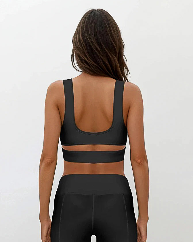 Eine Frau mit langen braunen Haaren blickt in die Ferne. Sie trägt das Main Design Flow Active Top mit einem breiten horizontalen Träger und dazu passende Leggings mit hoher Taille vor einem schlichten hellen Hintergrund.