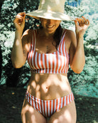 Eine Frau in einem Main Design Hale Bikini-Oberteil mit roten und weißen Streifen und Rundhalsausschnitt lächelt im Freien und hält ihren Strohhut. Das Sonnenlicht fällt durch die Bäume und das Grün im Hintergrund.