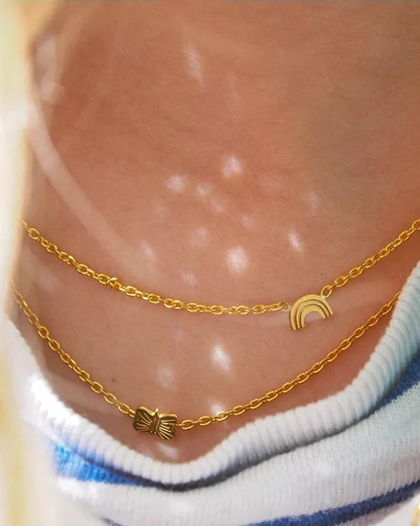 Nahaufnahme einer Person, die zwei zarte Adorabili Halsketten aus 24 Karat vergoldetem Gold - eine mit einem Schmetterlingsanhänger, eine mit einem Regenbogenanhänger - über einem blau-weiß gestreiften Hemd trägt. Das Sonnenlicht schimmert auf der Haut. Ideal als Geschenk für Kinder.