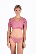 Eine Frau blickt vor einem weißen Hintergrund nach vorne und trägt das kurzarm Lycra-Crop-Top „Evie“ von Main Design und ein dazu passendes Bikinihöschen aus geripptem Stoff in Altrosa, beide aus recycelten Materialien für einen umweltfreundlichen Touch gefertigt.