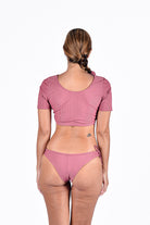 Eine Frau blickt von der Kamera weg und trägt das Main Design Evie Crop Top Kurzarm Lycra, einen gerippten, malvenfarbenen Zweiteiler aus recyceltem Material mit kurzen Ärmeln und seitlich gebundenen Hosenbeinen. Ihr Haar ist zu einem tiefen Zopf geflochten, der ein kleines Tattoo im unteren Rückenbereich freigibt.
