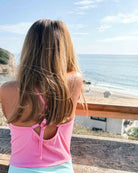 Ein Mädchen mit langen, hellbraunen Haaren trägt den Lena Mädchen Badeanzug von Main Design aus recycelten Materialien und steht an einem Holzgeländer mit Blick auf den Sandstrand und die Meereswellen unter blauem Himmel.