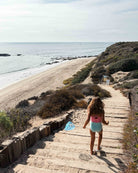 Ein Mädchen im Lena Mädchen Badeanzug von Main Design aus recycelten Materialien läuft über sandige Holzstufen zum ruhigen Strand, mit Meer, Felsenküste und bewachsenen Klippen unter klarem Himmel.