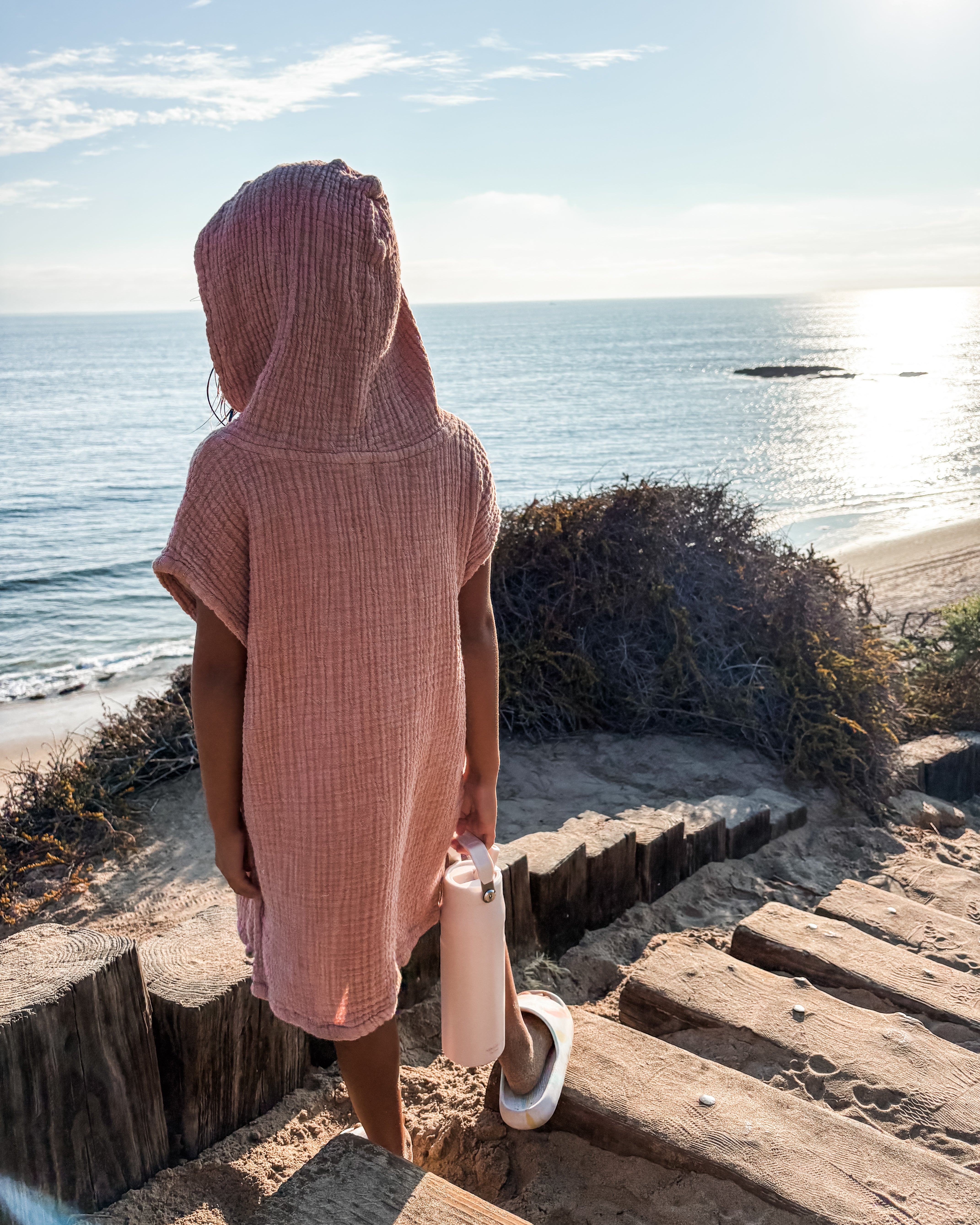 Ein Kind in einem rosafarbenen Kinder Badeponcho Flow von Main Design steht auf sandigen Holzstufen mit Blick auf den Ozean, hält eine Wasserflasche und Sandalen in der Hand, mit sonnenbeschienenen Wellen und Küstengebüsch im Hintergrund.