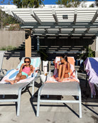 Zwei junge Mädchen in Badeanzügen entspannen sich am Pool und genießen ein Eis auf Liegestühlen, die mit Maxi Strandtuch De La Mer von Main Design bezogen sind. Bunte Handtücher und helles Sonnenlicht sorgen für einen verspielten Touch unter der Pergola.